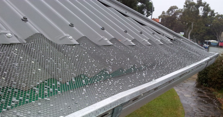 A photo shows non-combustible gutter leaf screens in place on the edge of a roof. The mesh stretches from the edge of the roof material to the outer lip of the gutter, preventing leaves and other debris from entering the guttering and downpipes.